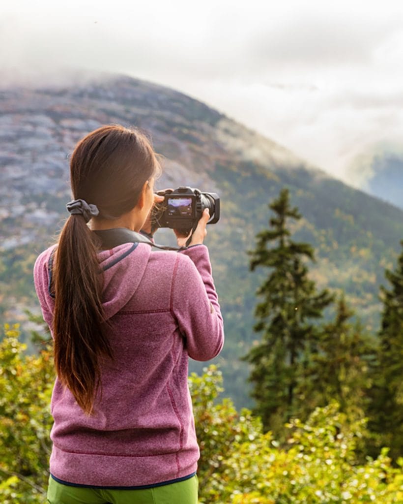 a woman holding a camera taking a photo of an alpine Alaskan landscape