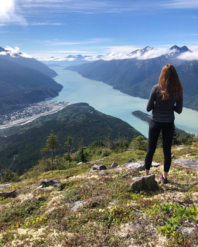 a woman on a high elevation landscape looking down at a bay and port town in Alaska